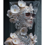 A surreal portrait of a model wearing an elaborate silver rhinestone face mask, smokey sunglasses. and matching embellished diamond collar necklace, decorated with delicate pink flowers, set against a dark grey background.