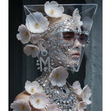 A surreal portrait of a model wearing an elaborate silver rhinestone face mask, smokey sunglasses. and matching embellished diamond collar necklace, decorated with delicate pink flowers, set against a dark grey background.