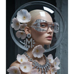 A fashion surreal portrait of a model with platinum blonde hair wearing oversized clear eyewear, diamond earrings with "M" monogram and matching diamond necklace. The model is adorned with pale pink crystal flowers with her head encapsulated with a transparent crystal class dome. The scene is set against a dark textured background. Artwork by MizDragonfly.