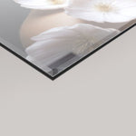 A close-up of a 4mm rectangular glass panel or mirror with a glossy finish, showing a reflection of white flowers and a soft beige background. The glass has a thin dark edge and is photographed at an angle against a light gray background.