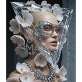 A surreal portrait of a model with platinum blonde hair wears a large, clear faceted diamond-like geometric headpiece, silver rhinestone eyewear, a matching diamond necklace, and a large diamond 'M' earring, surrounded by white flowers against a dark grey background.
