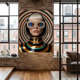 A large canvas wall art print featuring a stylized portrait of a woman with silver hair, rainbow-striped sunglasses, and a black, gold and turquoise striped top, displayed on a brick wall in a modern loft room with wood floors and large windows.