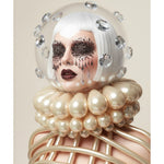A surreal fashion portrait of a person with a white bob hair style, dramatic black glitter eye masks, and dark sparking red lipstick, wearing a clear dome helmet with diamonds and a fluted pearl-like ruff collar, with metallic gold cage-like ribbing around the neck and shoulders, set against a warm beige background.