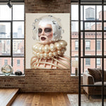 A large vertical wall art displayed in a modern loft-style room with exposed brick walls, wood floors, and large windows. The artwork depicts a surreal woman with a clear glass helmet, pale face, dark crystal eye patches with crystal tears, and a voluminous pearl-like collar and ribbed body against a light beige background.