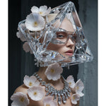 A surreal portrait of a model wearing a sculptural clear crystal geometric headpiece with white flowers, a matching crystal necklace, and clear eyeglasses against a dark gray background.