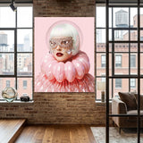 A large vertical wall art featuring a stylized portrait of a woman with a pink bubble helmet and voluminous pink ruffled clothing, displayed on a brick wall in a modern loft room with wood floors and large windows.