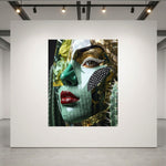 A large square art print depicting a stylized close-up portrait of a woman's face with green and gold textures, red lips, and black diamond cheek detail, displayed on a white gallery wall in a modern gallery space with track lighting. 