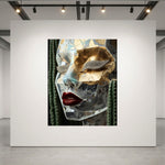 A large framed art print displayed on a white gallery wall, featuring a stylized close-up portrait of a women's face wearing a reflective metallic mask with gold, silver, and red details, set against a white wall in a modern gallery space.