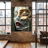 A large canvas wall art print featuring a stylized, transparent acrylic face with gold, silver, and red details, set in a cacti decor, displayed on a brick wall in a modern loft-style room with wood floors and windows.