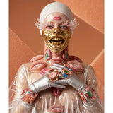 A person wearing a white head covering, a gold face mask, silver gloves, and a clear plastic jacket, with multiple pink lips attached or styled on the face, neck, and chest, posed against a peach-colored textured background. 