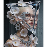 A fashion editorial portrait of a model wearing a large triangular clear crystal frame over her face, paired with a diamond necklace and earrings with "M" monogram, decorated with pale pink flowers against a dark grey background. Limited edition surreal artwork by MizDragonfly.