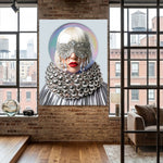A large vertical wall art print featuring a stylized portrait of a woman with silver eye masks, a voluminous silver collar, and white hair, displayed on a brick wall in a modern loft room with wood floors and windows.