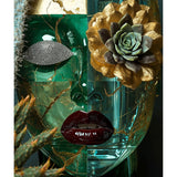 A decorative sculptural face made from translucent teal glass with gold marbling, a textured metallic diamond eye, glossy dark red lips, and a large gold-and-green succulent on the eye lip and cheek, shown against a dark cacti background. 